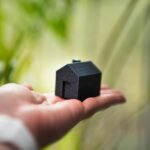 Close-up of a hand holding a miniature black house with greenery in the background.